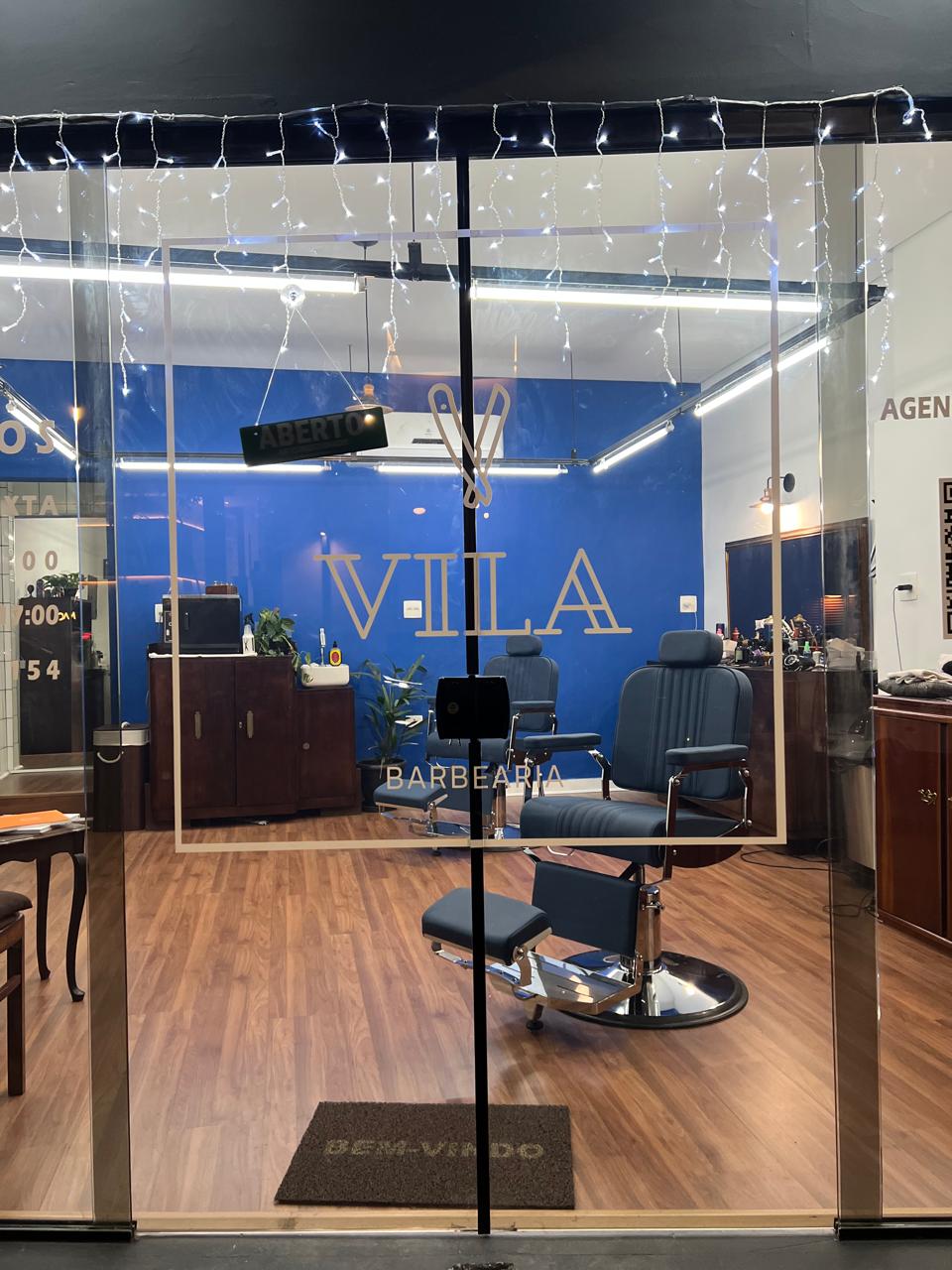 Porta de vidro da Vila Barbearia, com o logo pintado em dourado sobre a parede azul